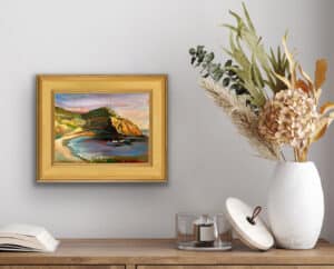 Original oil painting "Crystal Cove Sunset Cliffs" by Ron Reekers displayed in a traditional gold frame above a wooden console beside a white vase filled with dried botanical arrangements.