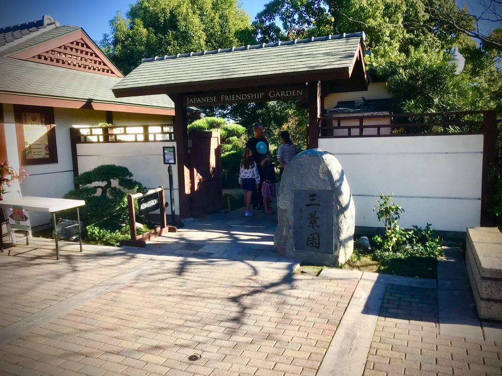 Traditional Japanese-style entrance to the Japanese Friendship Garden & Museum in Balboa Park, San Diego, with visitors walking through the wooden gate and a stone monument in the foreground.